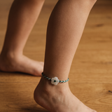 A close-up of a child's bare feet standing on a wooden floor, wearing a delicate braided anklet with a white flower charm.