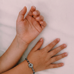 A close-up of two hands resting on a light pink fabric surface, with one hand wearing a silver-toned charm bracelet featuring a central white stone.