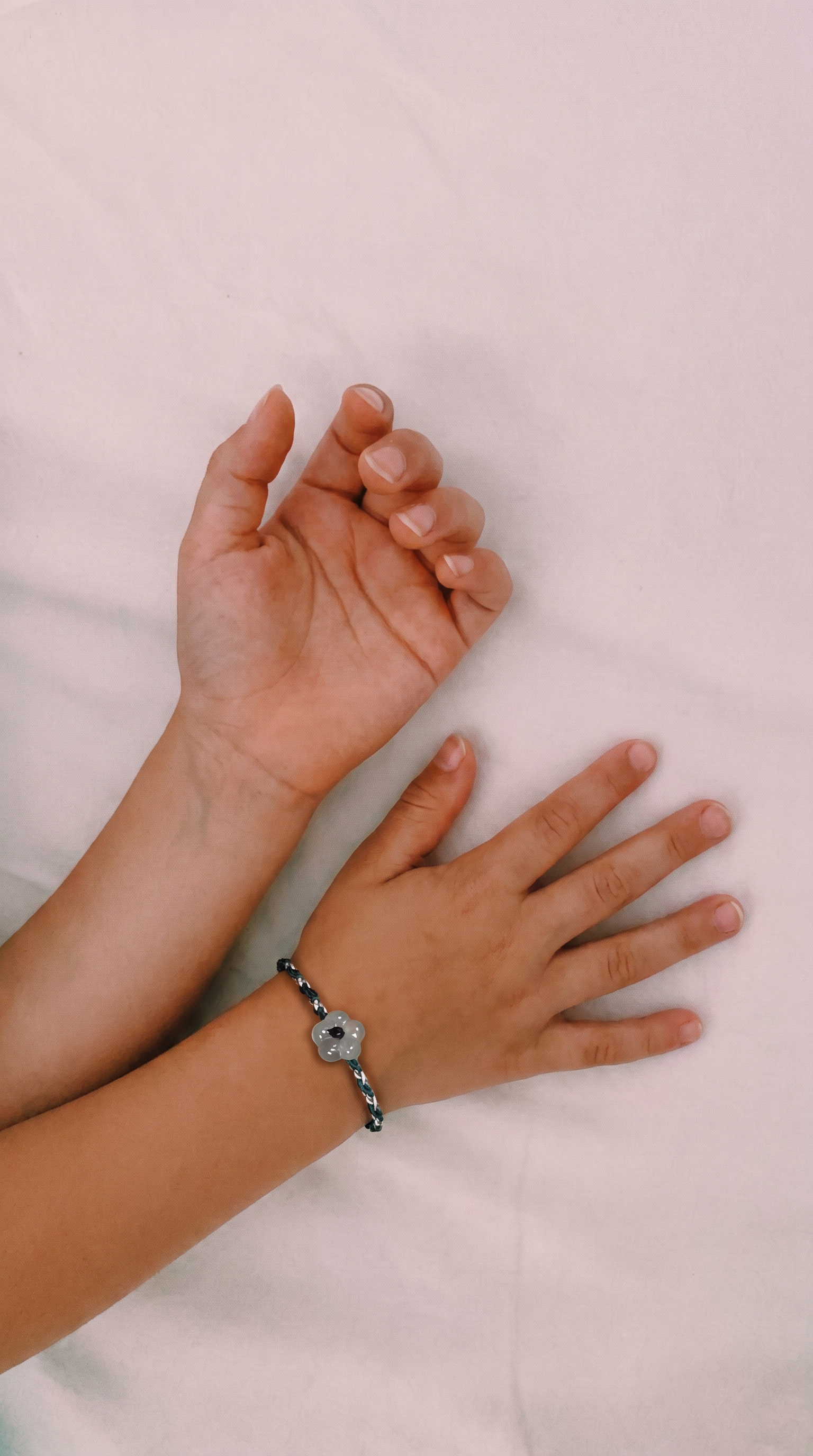 A close-up of two hands resting on a light pink fabric surface, with one hand wearing a silver-toned charm bracelet featuring a central white stone.