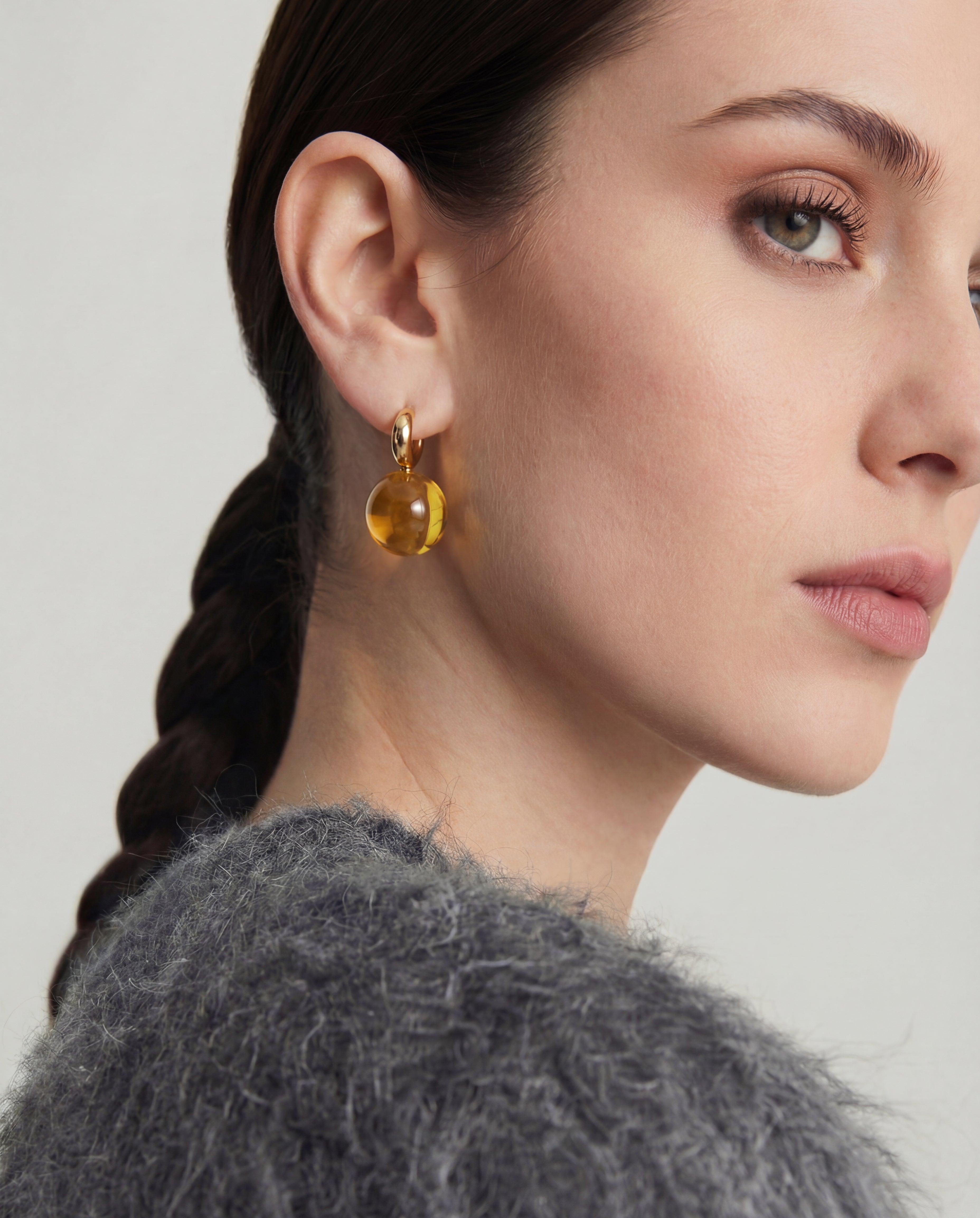 Close-up of a woman wearing a gold gemstone earring with a gray fur collar on a light background