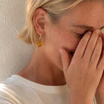 A close-up lifestyle shot of a woman with blonde hair covering her face with her hands, wearing a small gold hoop earring with a round amber-yellow drop pendant against a textured beige wall.
