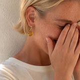 A close-up lifestyle shot of a woman with blonde hair covering her face with her hands, wearing a small gold hoop earring with a round amber-yellow drop pendant against a textured beige wall.