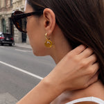 A woman in profile wearing a gold huggie hoop earring with a translucent amber-yellow circular drop pendant, photographed outdoors on a city street.