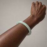A light green jadeite bangle bracelet worn on a person's wrist, shown against a plain neutral background.