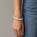 A person wearing a smooth, light green jadeite bangle bracelet, paired with blue denim jeans, shown from the waist down against a neutral gray background.