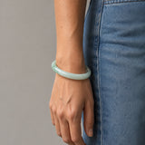 A person wearing a smooth, light green jadeite bangle bracelet, paired with blue denim jeans, shown from the waist down against a neutral gray background.