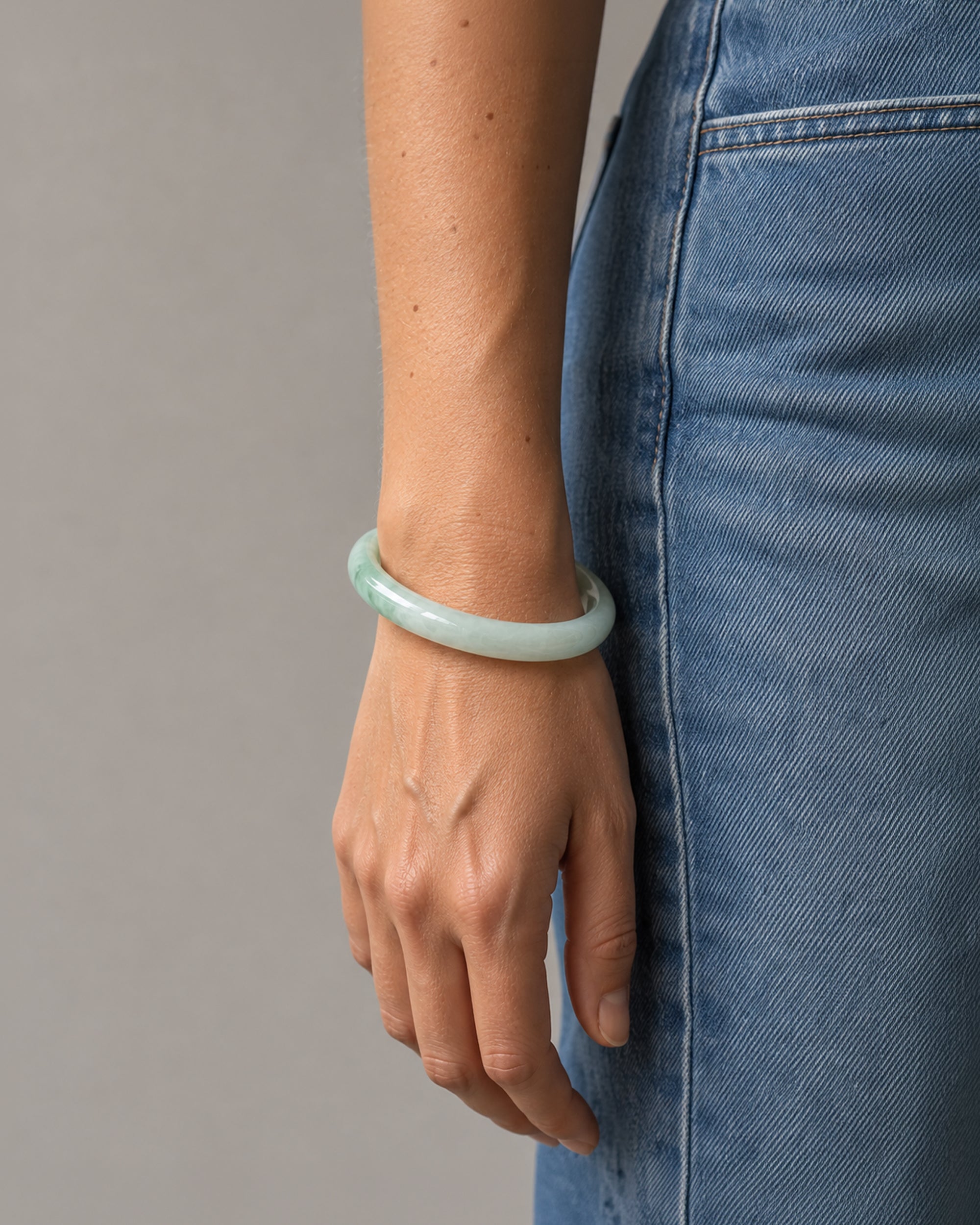 A person wearing a smooth, light green jadeite bangle bracelet, paired with blue denim jeans, shown from the waist down against a neutral gray background.