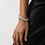 A light green jadeite bangle bracelet worn on a person's wrist, shown against a black satin dress and a plain light gray background.