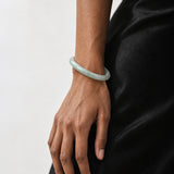 A light green jadeite bangle bracelet worn on a person's wrist, shown against a black satin dress and a plain light gray background.