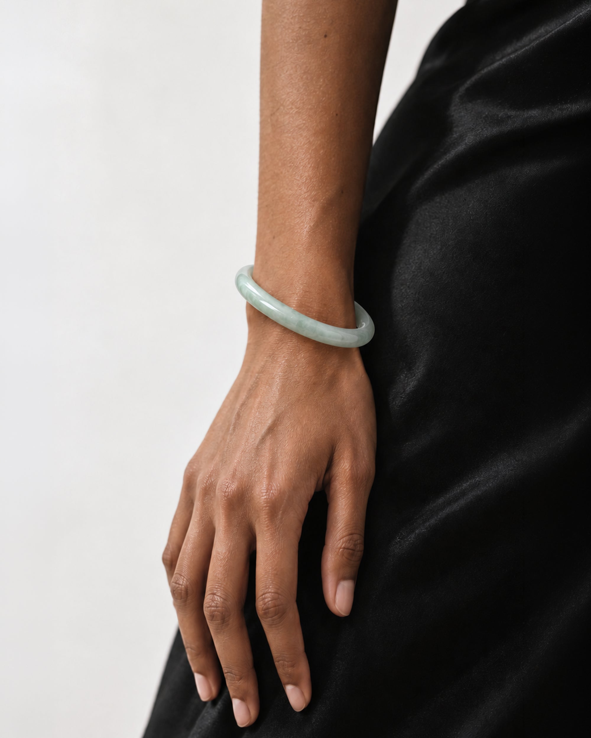 A light green jadeite bangle bracelet worn on a person's wrist, shown against a black satin dress and a plain light gray background.