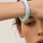 A close-up of a woman's wrist wearing a smooth, semi-translucent jadeite bangle with a pale green and lavender-blue gradient.