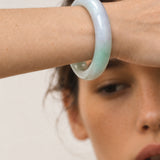 A close-up of a woman's wrist wearing a smooth, semi-translucent jadeite bangle with a pale green and lavender-blue gradient.