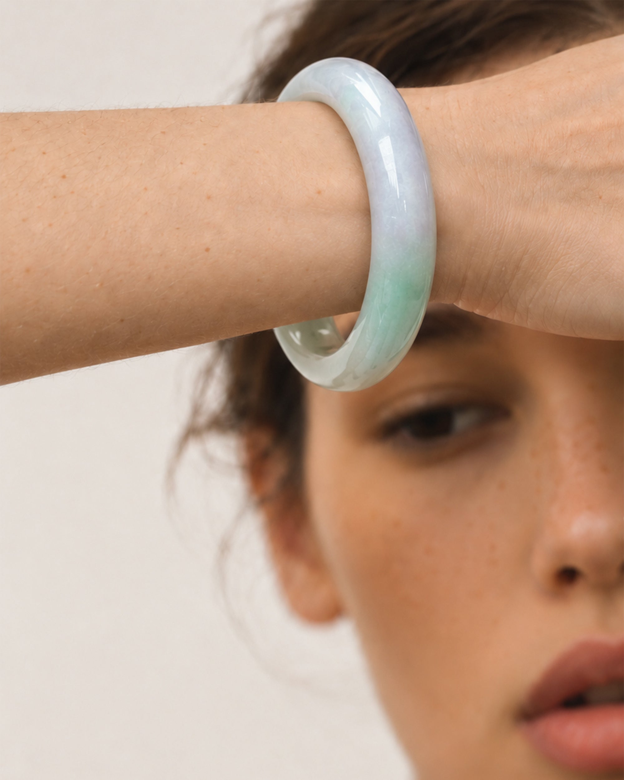 A close-up of a woman's wrist wearing a smooth, semi-translucent jadeite bangle with a pale green and lavender-blue gradient.