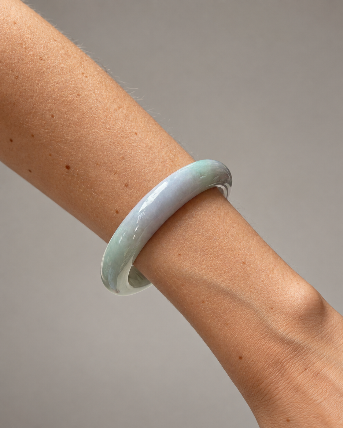 A light green and lavender jadeite bangle bracelet worn on a person's wrist, shown against a neutral gray background.
