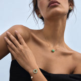 A close-up lifestyle shot of a model wearing a gold jewelry set featuring a delicate necklace with a small green stone, a matching bracelet with a green ring and pearl, and earrings, set against a light blue sky background.