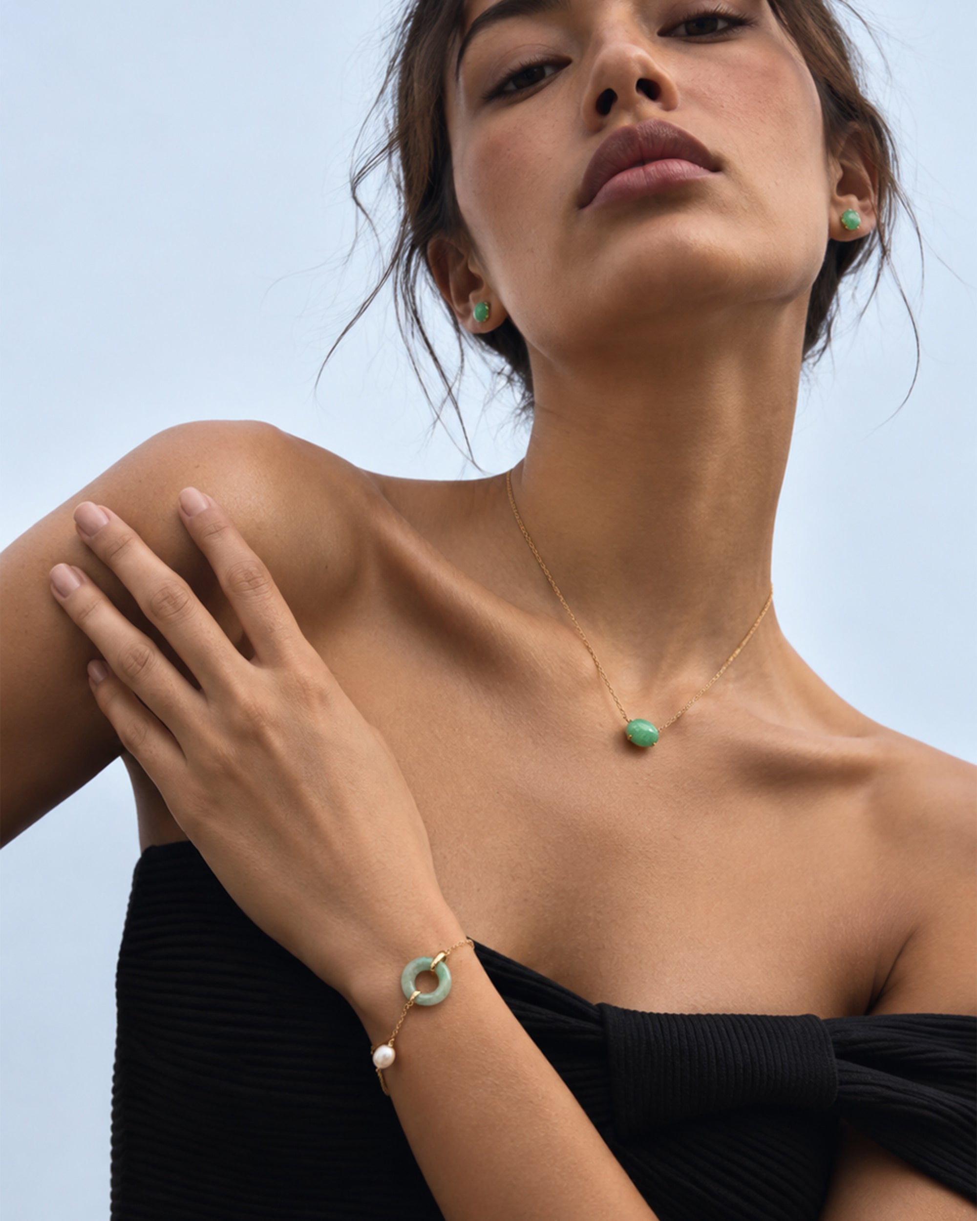 A close-up lifestyle shot of a model wearing a gold jewelry set featuring a delicate necklace with a small green stone, a matching bracelet with a green ring and pearl, and earrings, set against a light blue sky background.