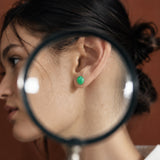 A close-up lifestyle shot of a woman wearing a small green stud earring, viewed through a magnifying glass held in front of her ear. 