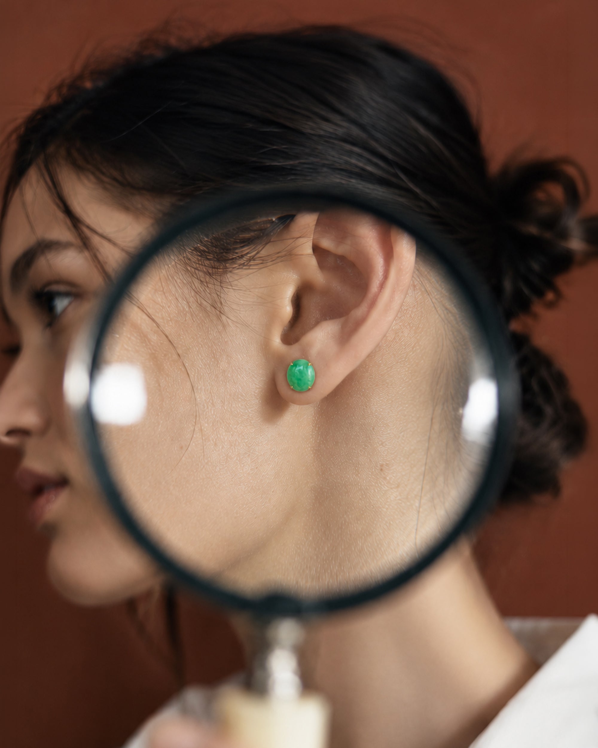 A close-up lifestyle shot of a woman wearing a small green stud earring, viewed through a magnifying glass held in front of her ear. 