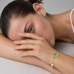 A close-up lifestyle shot of a model resting her head on her arms, showcasing a delicate gold chain bracelet with a light green jade-like ring charm, a small white pearl, and a gold earring.