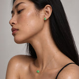 A close-up side profile of a woman wearing a delicate gold necklace and matching stud earring, both featuring a small round green cabochon stone, against a neutral gray background.