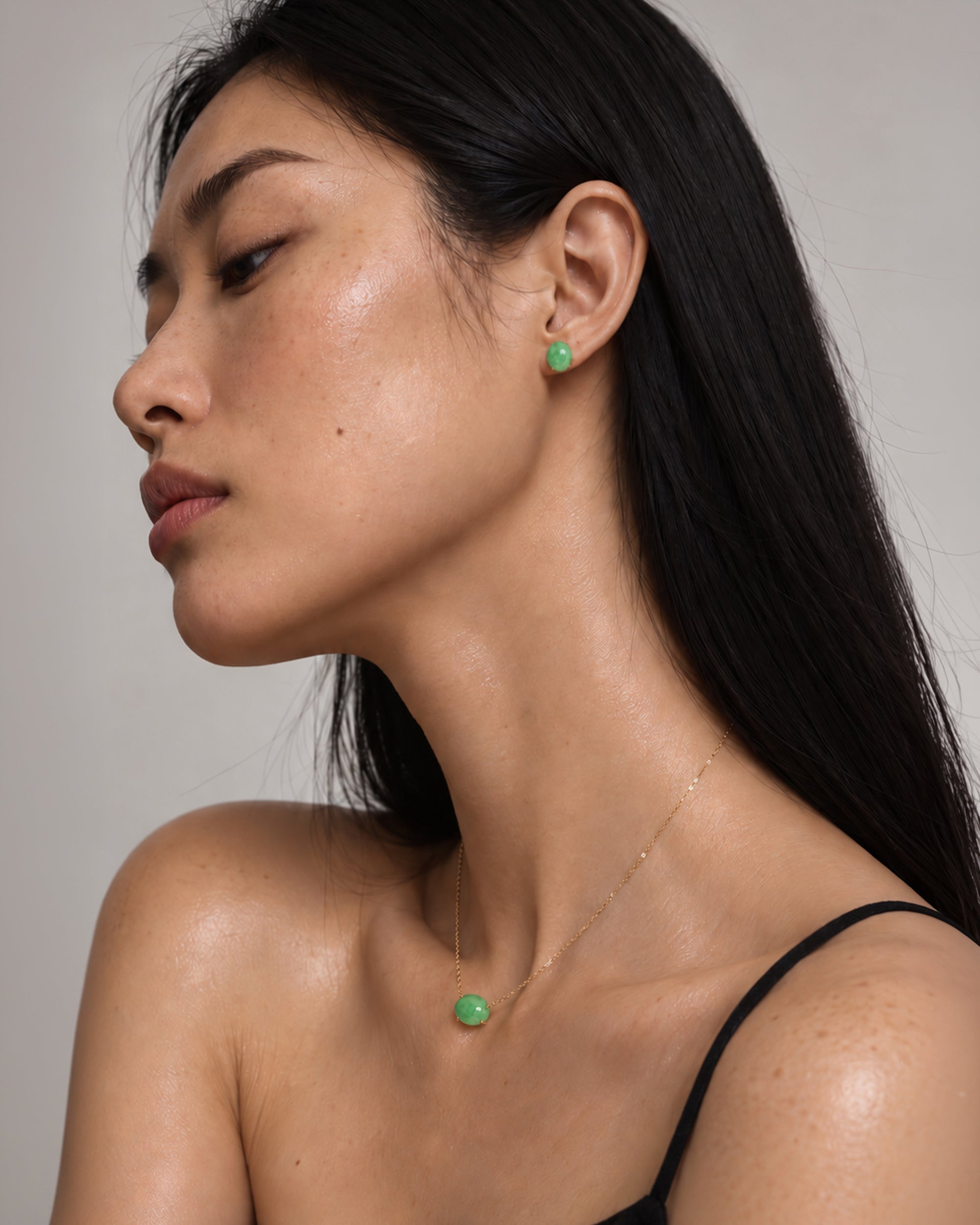 A close-up side profile of a woman wearing a delicate gold necklace and matching stud earring, both featuring a small round green cabochon stone, against a neutral gray background.