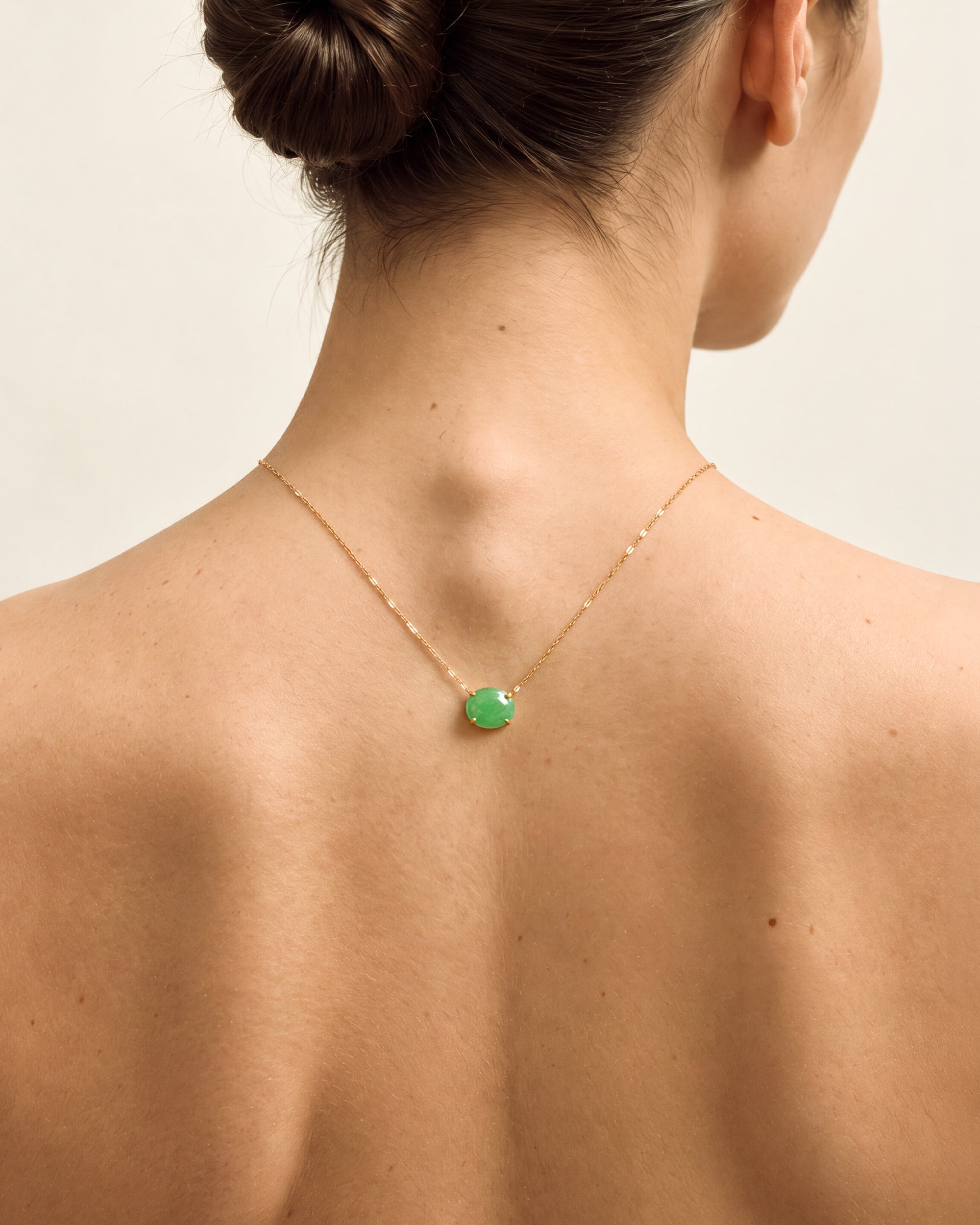 A woman shown from the back wearing a delicate gold chain necklace with a small round green pendant against a plain light beige background.