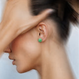A close-up side profile of a woman wearing a small round green stud earring, with her hand partially covering her face against a soft light background.