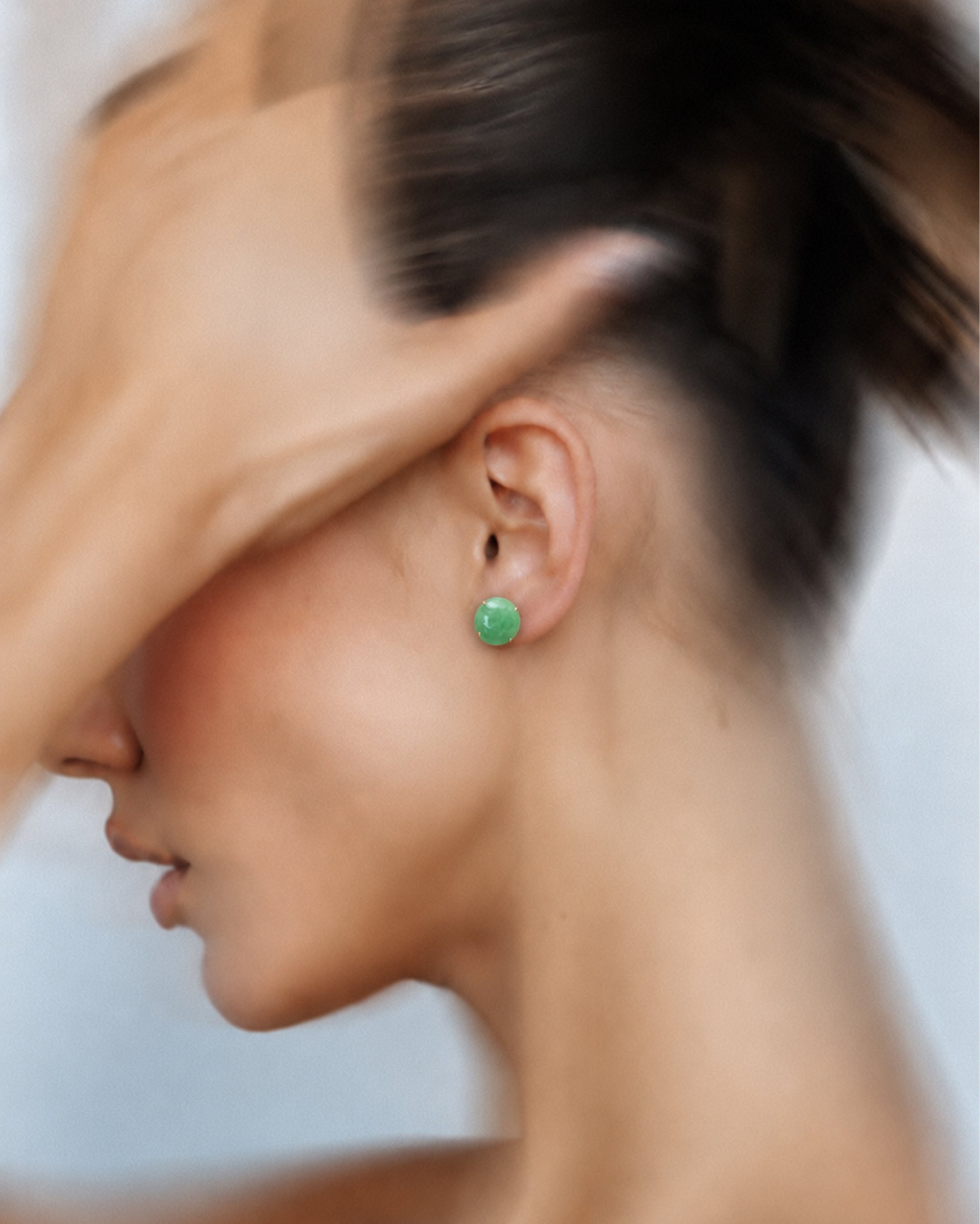 A close-up side profile of a woman wearing a small round green stud earring, with her hand partially covering her face against a soft light background.