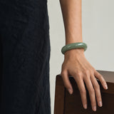 A person wearing a smooth, glossy light green jadeite bangle bracelet, shown from the waist down with their hand resting on a wooden surface.