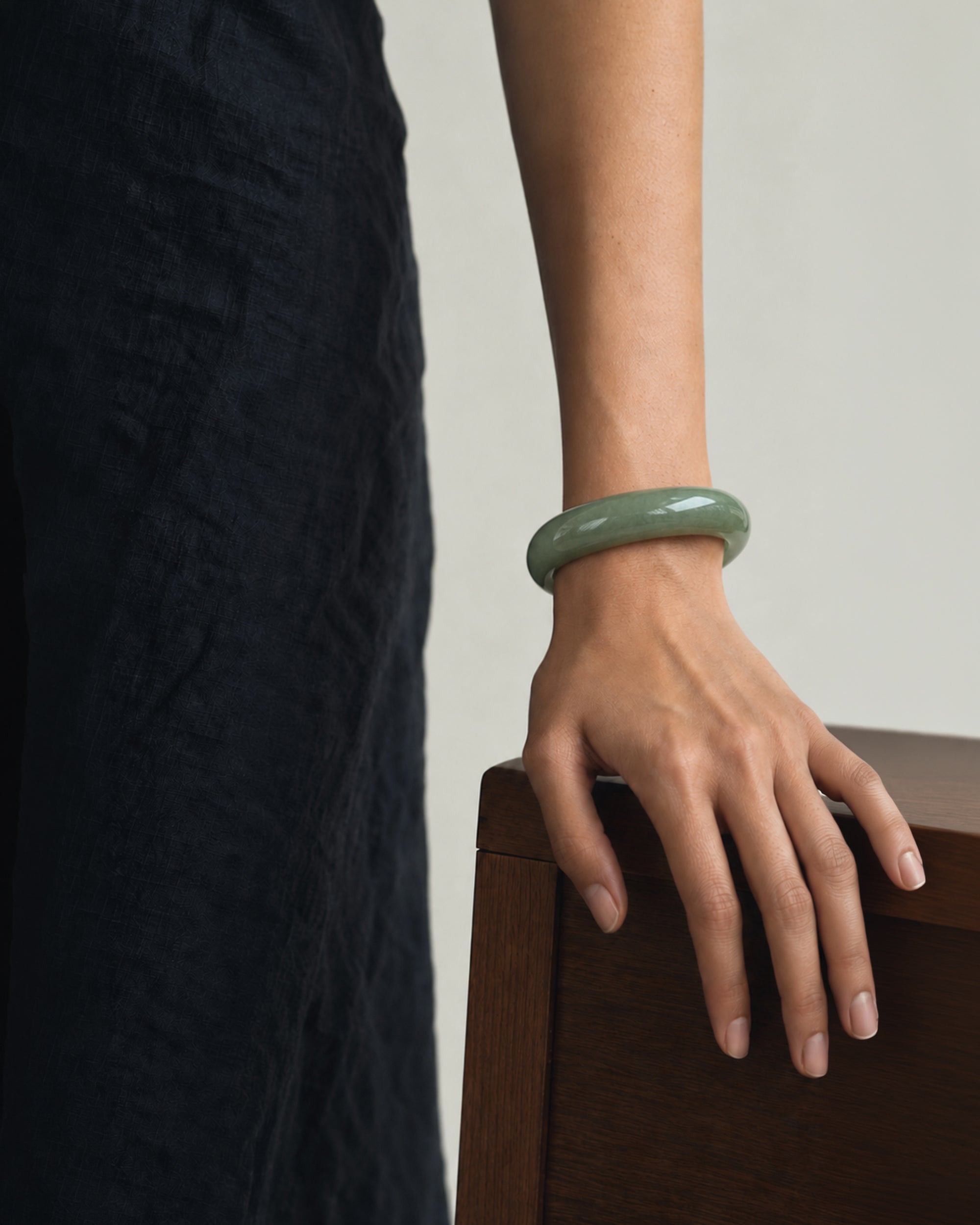 A person wearing a smooth, glossy light green jadeite bangle bracelet, shown from the waist down with their hand resting on a wooden surface.