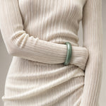 A close-up of a person wearing a cream-colored, long-sleeve pleated ribbed top and a smooth, light green jade bangle bracelet on the wrist.