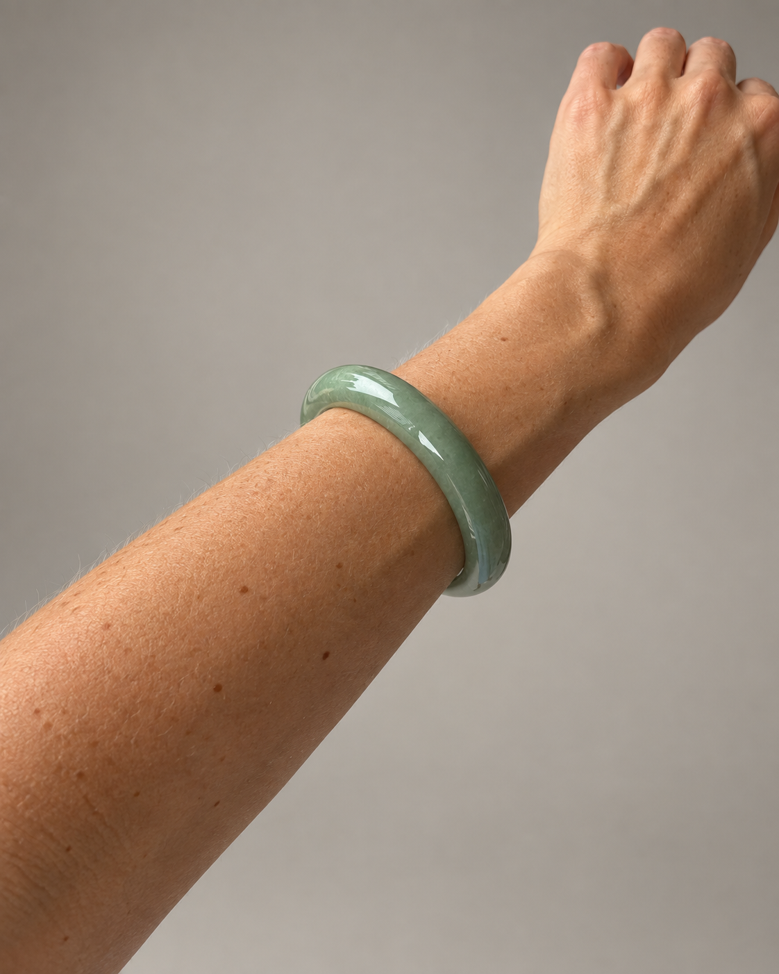 A light green jadeite bangle bracelet worn on a person's wrist, shown against a plain gray background.