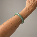 A light green jadeite bangle bracelet worn on a person's wrist, shown against a plain gray background.