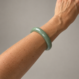A light green jadeite bangle bracelet worn on a person's wrist, shown against a plain gray background.