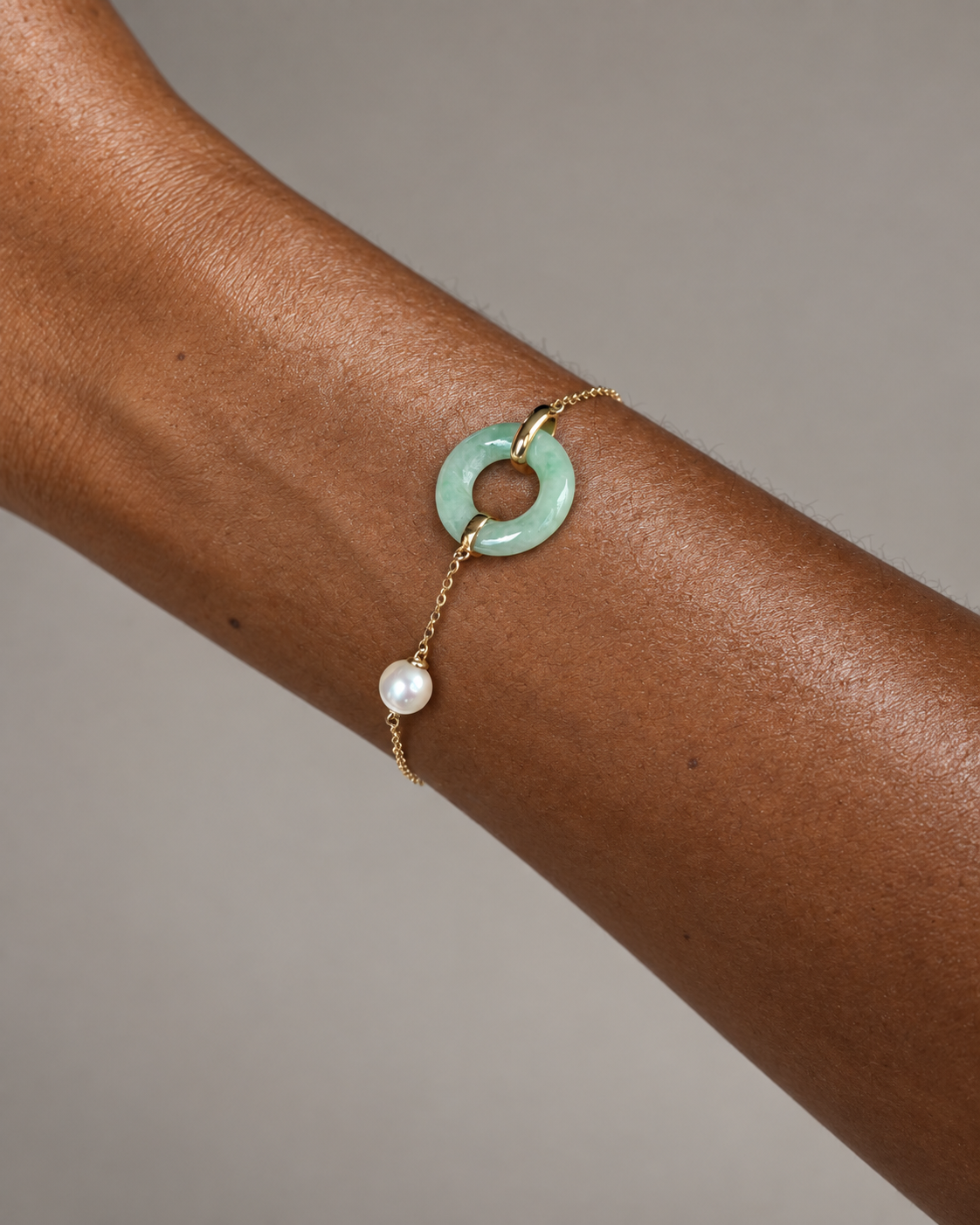 A delicate gold chain bracelet worn on a person's wrist, featuring a light green jadeite ring charm and a dangling white pearl bead, set against a neutral gray background.
