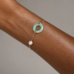 A delicate gold chain bracelet worn on a person's wrist, featuring a light green jadeite ring charm and a dangling white pearl bead, set against a neutral gray background.