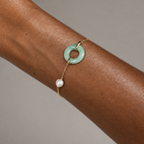 A delicate gold chain bracelet worn on a person's wrist, featuring a light green jadeite ring charm and a dangling white pearl bead, set against a neutral gray background.