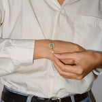 A close-up of a person wearing a delicate gold chain bracelet with a light green jade disc and a white pearl, shown on their wrist while they adjust it with their fingers.