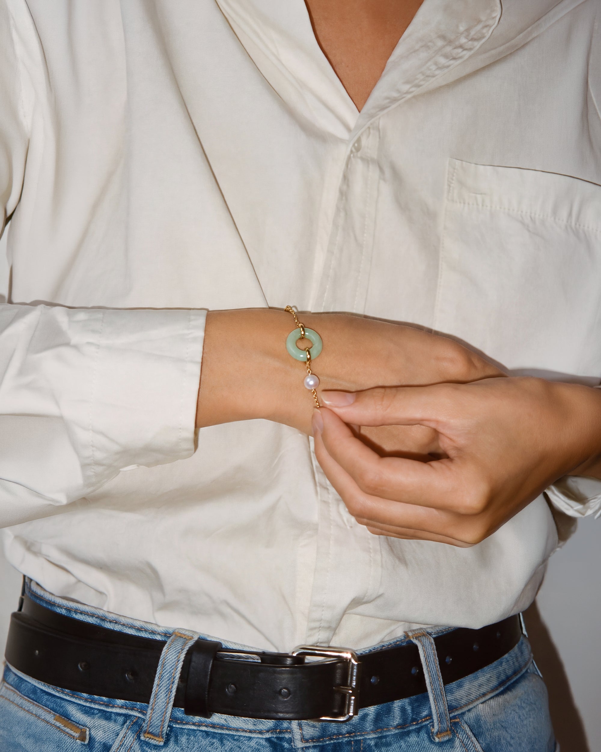 A close-up of a person wearing a delicate gold chain bracelet with a light green jade disc and a white pearl, shown on their wrist while they adjust it with their fingers.