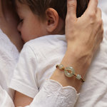 A close-up lifestyle shot of a mother holding and kissing her baby, with the baby wearing a white dress. The mother's hand is adorned with a delicate gold bracelet featuring light green jade-like round beads.
