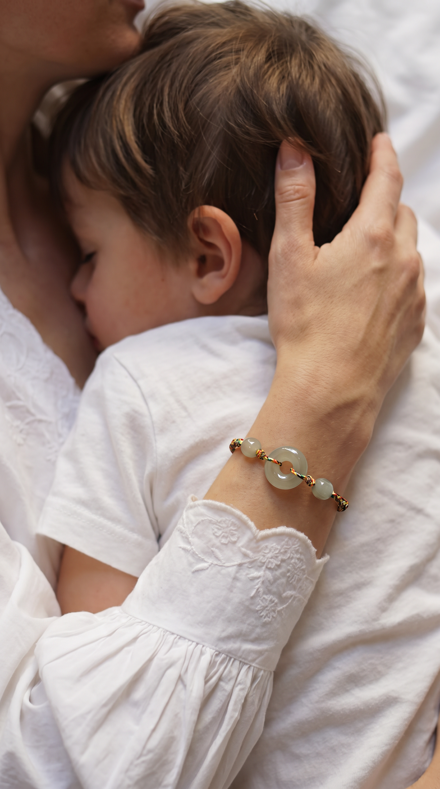 A close-up lifestyle shot of a mother holding and kissing her baby, with the baby wearing a white dress. The mother's hand is adorned with a delicate gold bracelet featuring light green jade-like round beads.