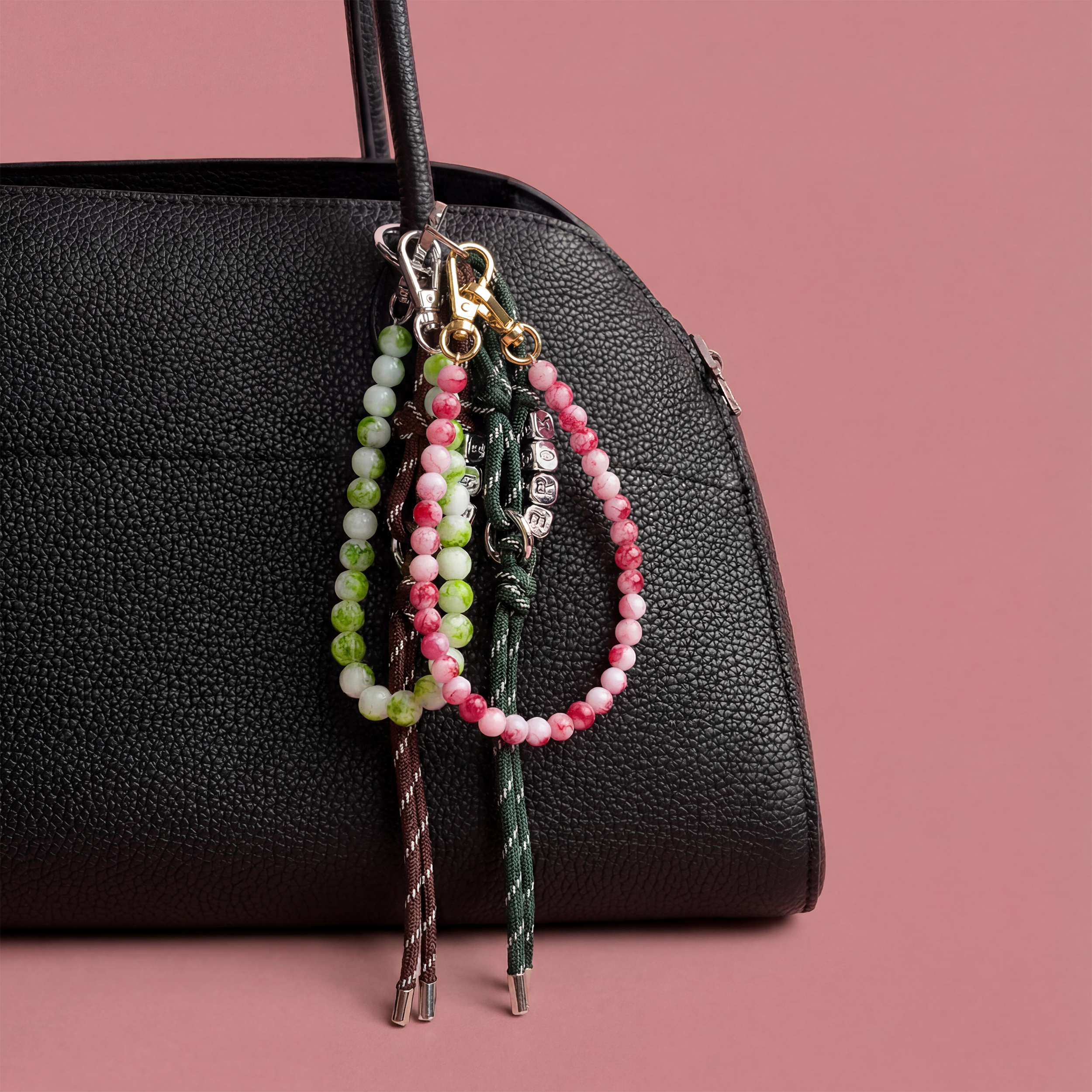Three beaded keychains hanging from a black handbag against a pink background