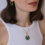 Woman wearing a gold necklace with a green pendant against a neutral background
