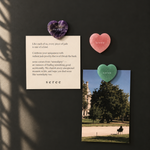 Three heart-shaped jade pendants in purple, pink, and green are displayed on a dark wall alongside a cream card with a quote and a printed photo. The brand name 'seree' is visible on the hearts and the card.