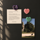 Three heart-shaped jade pendants in purple, pink, and green are displayed on a dark wall alongside a cream card with a quote and a printed photo. The brand name 'seree' is visible on the hearts and the card.