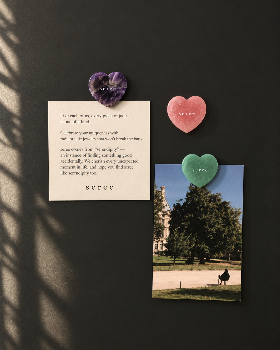 Three heart-shaped jade pendants in purple, pink, and green are displayed on a dark wall alongside a cream card with a quote and a printed photo. The brand name 'seree' is visible on the hearts and the card.