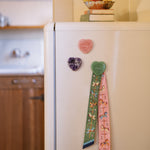 Three heart-shaped crystal magnets in pink, purple, and green on a white refrigerator. The green magnet is holding a decorative patterned twilly.