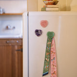 Three heart-shaped crystal magnets in pink, purple, and green on a white refrigerator. The green magnet is holding a decorative patterned twilly.