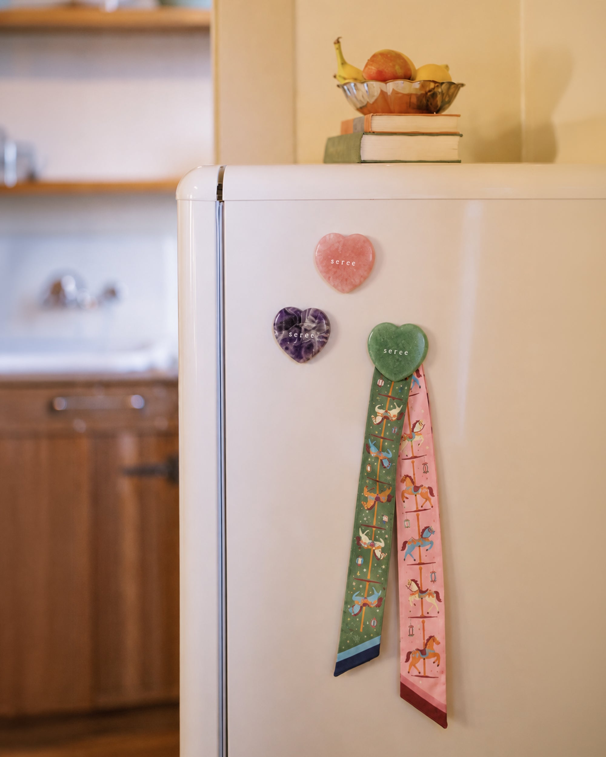 Three heart-shaped crystal magnets in pink, purple, and green on a white refrigerator. The green magnet is holding a decorative patterned twilly.