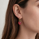 Close-up of a person wearing a gold earring with a red strawberry gemstone against a neutral background